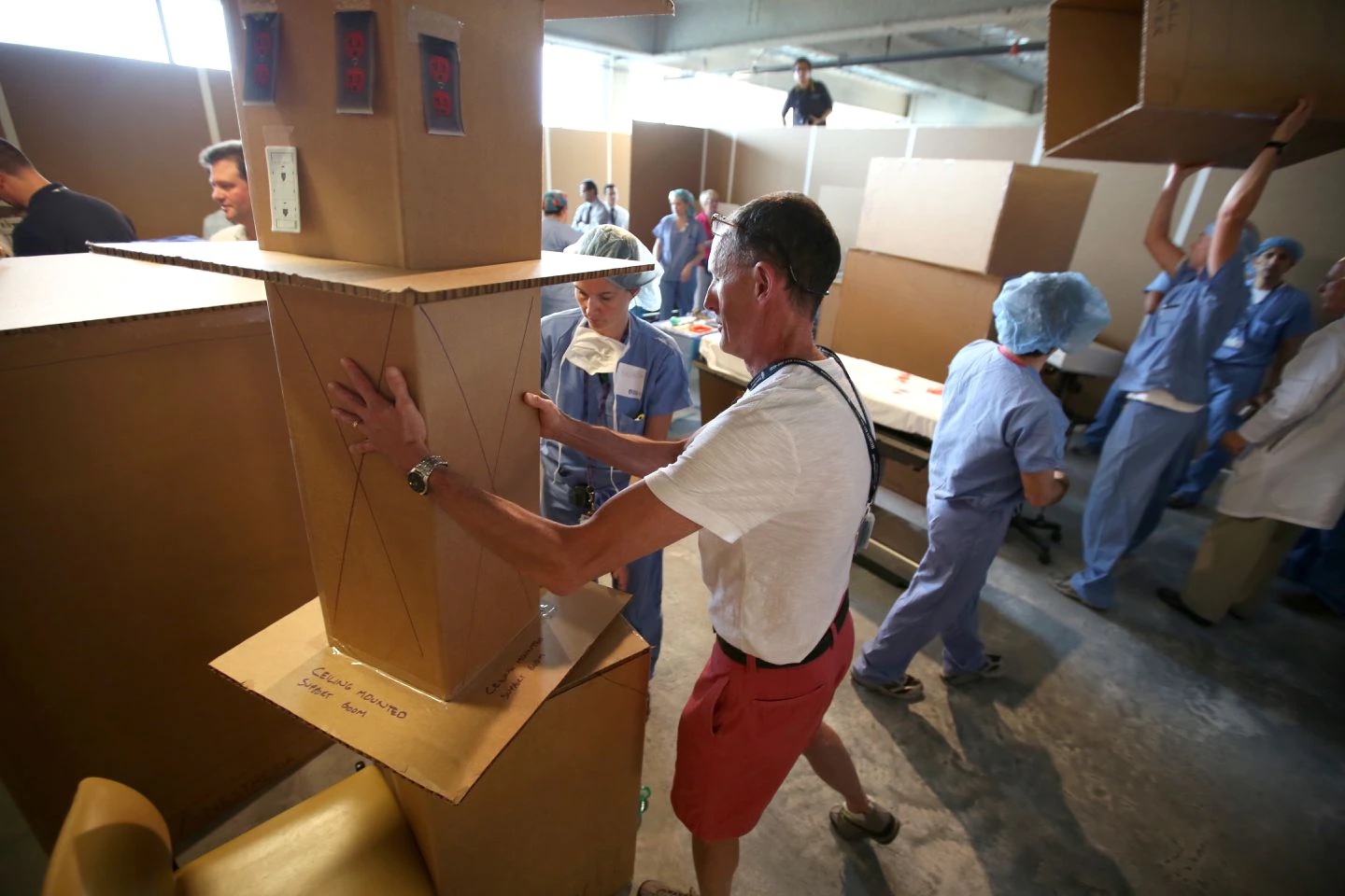 穿着蓝色手术服的医生和一名穿红色短裤和白衬衫的男子都在房间里移动纸板箱，用它们来建造大楼。